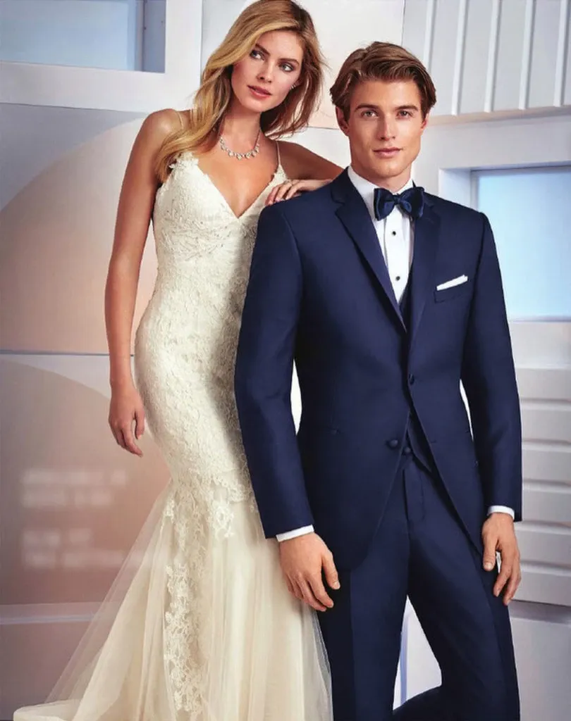 A bride and groom are stylishly posing indoors. The groom wears a classic navy blue tuxedo by Jaco Uomo, perfectly tailored for a wedding, featuring a crisp white shirt, dark bow tie, and pocket square. The elegant attire emphasizes sophistication, suitable for a formal wedding setting. The bride is in a sleeveless lace wedding gown with a plunging neckline and long train, accessorized with a delicate necklace. This image reflects a sophisticated wedding style, ideal for showcasing wedding tuxedo options from Jaco Uomo for Mariage Elegant, Canada.