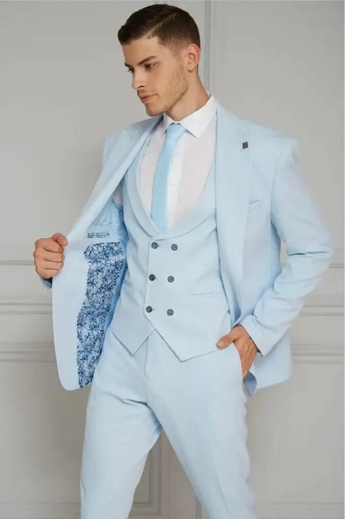 The image shows a groom wearing a tailored light blue tuxedo from Jaco Uomo, categorized under Wedding Tuxedos. The ensemble includes a crisp white dress shirt, a matching light blue tie, and a double-breasted waistcoat, creating a stylish and coordinated look. The groom is subtly displaying the inside of the jacket, which features an elegant floral lining, adding a touch of sophistication. This outfit is ideal for a modern, fashionable wedding, offering a fresh alternative to traditional black tuxedos.