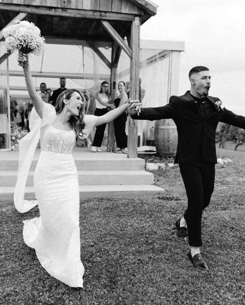 A joyful couple is walking hand-in-hand outside, showcasing their wedding attire. The groom is wearing a sleek tuxedo from M/2 Boutiques, fitting for the Wedding Tuxedo category, complementing his polished look. The bride, exuberant in her elegant white wedding gown, holds a bouquet of flowers. They are both smiling broadly, exuding excitement and happiness. In the background, family and friends appear to celebrate under a rustic wooden structure, adding to the festive wedding atmosphere. The scene captures a moment of delight during the couple's special day, reflecting elegance and joyful celebration.