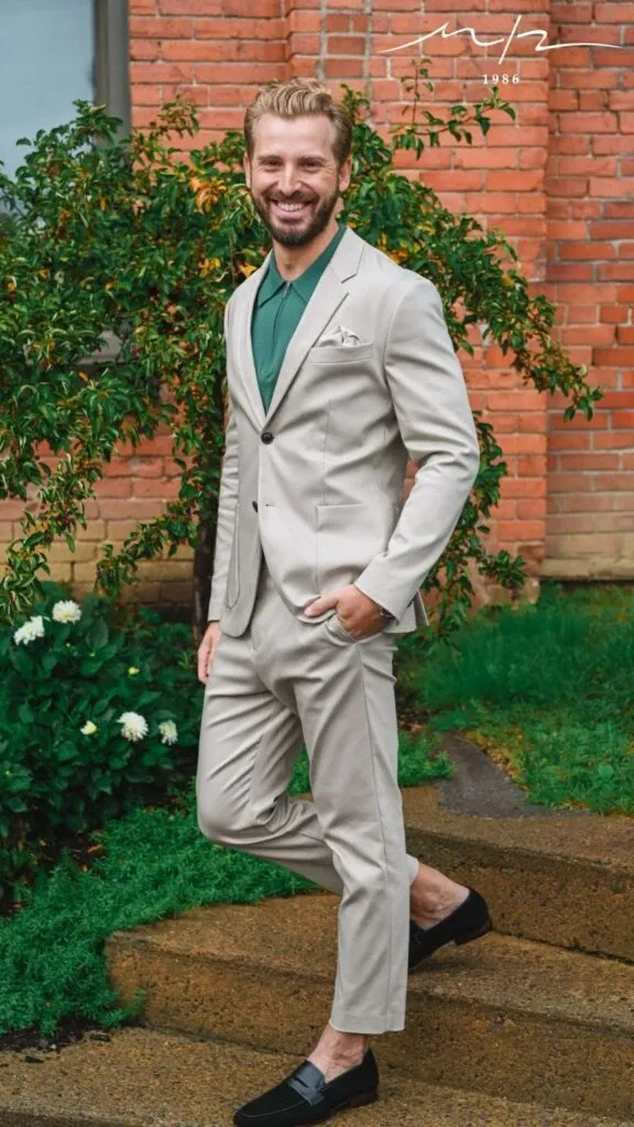 A groom is elegantly dressed in a light beige tuxedo by M/2 Boutiques, a vendor in the Wedding Tuxedo category. The tuxedo is paired with a green dress shirt and black loafers, creating a modern and stylish look suitable for weddings. The groom stands confidently in front of a brick wall with lush greenery in the background, showcasing the sophistication and quality of the attire offered by Mariage Elegant in Canada.