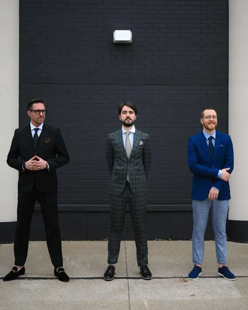 Three men stand in front of a dark brick wall, each showcasing different styles of wedding tuxedos from M/2 Boutiques under the Wedding Tuxedo category. The man on the left wears a classic black tuxedo with a white shirt and loafers, accentuated by a pocket square. The center man sports a gray checkered suit paired with a light blue shirt, looking polished and sophisticated. On the right, the man is dressed in a vibrant blue blazer with light gray pants, offering a modern twist to traditional wedding attire. These varied tuxedo styles highlight the versatility and elegance offered by M/2 Boutiques for weddings in Canada.