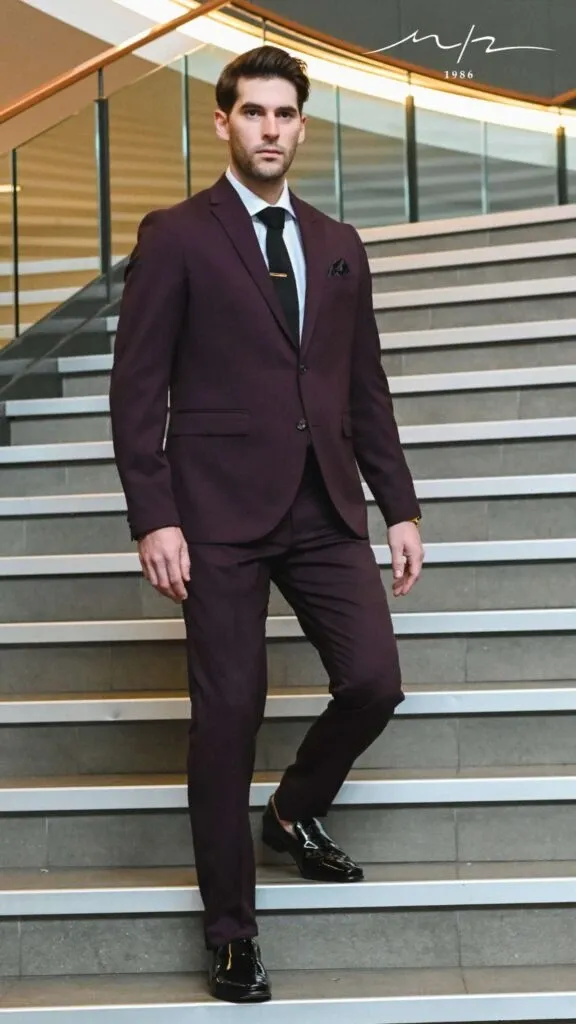 A man confidently descends a staircase wearing an elegant, deep burgundy tuxedo from M/2 Boutiques, featured in the Wedding Tuxedo category of Mariage Elegant, Canada. The tuxedo is beautifully tailored, complemented by a crisp white shirt, a sophisticated black tie, and a pocket square, perfect for a modern wedding. The man's polished black dress shoes complete the ensemble, highlighting the boutique's attention to detail and style. The setting, with its sleek, contemporary design, underscores the sophisticated appeal of M/2 Boutiques as a premier vendor for wedding attire.