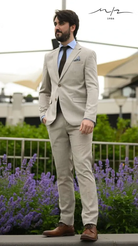 A groom is standing outdoors wearing a sophisticated beige wedding tuxedo from M/2 Boutiques, fitting the wedding tuxedo category. The tuxedo features a sleek design with a matching waistcoat, blue pocket square, and a dark tie. The groom poses against a backdrop of lush greenery and vibrant purple flowers, providing an elegant setting. This stylish attire from Mariage Elegant, Canada, exemplifies modern wedding fashion.