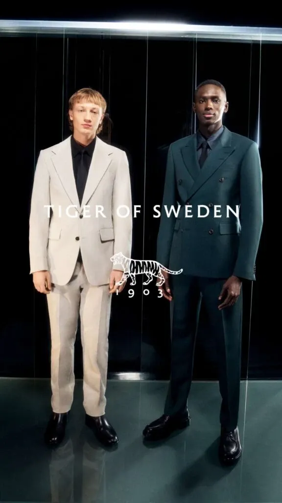 Two elegant gentlemen are standing side by side, showcasing wedding tuxedos from M/2 Boutiques, a vendor featured on Mariage Elegant under the Wedding Tuxedo category. The man on the left is dressed in a light grey tuxedo with a crisp black shirt, exuding modern sophistication. Meanwhile, the man on the right is wearing a dark teal double-breasted tuxedo paired with a matching shirt, adding a bold and classy touch to wedding attire options. Both tuxedos reflect high-quality craftsmanship and contemporary style suited for a stylish groom.
