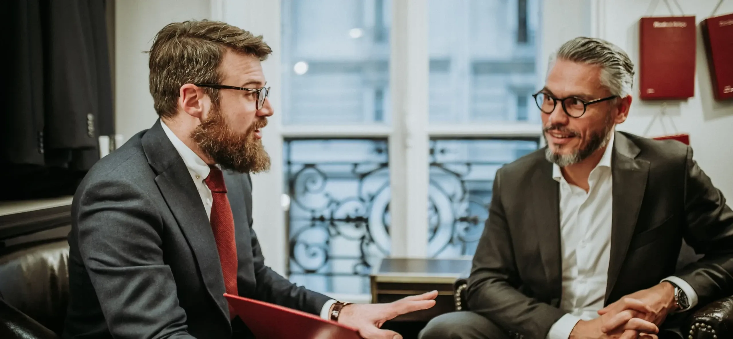 Two men are seated indoors, engaged in a discussion. They are both wearing formal wedding tuxedos, showcasing attire from the wedding vendor Blandin & Delloye, a specialist in elegant wedding tuxedos. The man on the left has a beard and is wearing glasses, a dark suit, and a red tie, while holding a red folder. The man on the right has neatly styled hair, a beard, and is also wearing glasses, along with a dark suit and a white shirt. The background features large windows with an intricate wrought iron design, adding to the sophisticated ambiance typical of a professional wedding planning setting.