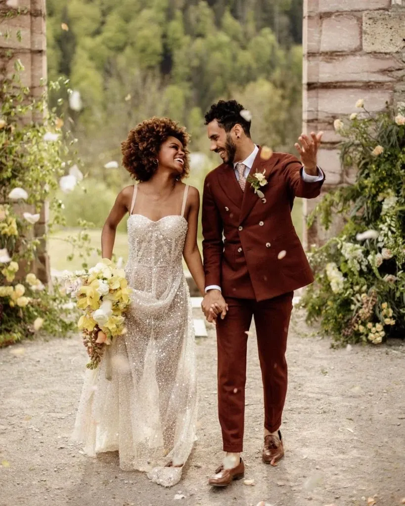 A joyful couple is walking hand-in-hand outdoors, celebrating their wedding. The groom is wearing a stylish, rust-colored suit with a boutonniere, provided by Blandin & Delloye under their wedding tuxedo category. The bride is dressed in a shimmering, semi-sheer gown, holding a bouquet of yellow and white flowers. They are surrounded by lush greenery and stone archways, creating a romantic and enchanting atmosphere. The natural background with trees enhances the elegance and charm of the moment, reflecting a perfect blend of style and nature for a memorable wedding day.