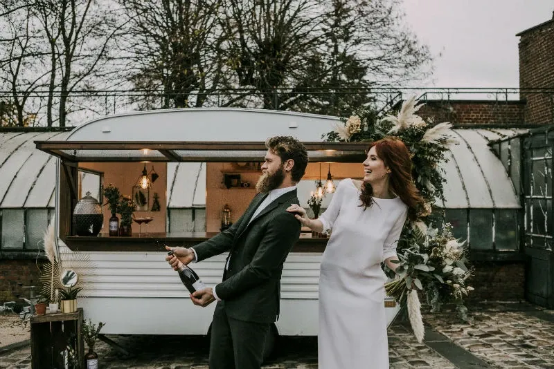 A groom in a stylish tuxedo by Blandin & Delloye, part of the wedding tuxedo category, playfully opens a bottle of champagne next to a bride in an elegant white dress. They stand in front of a vintage mobile bar adorned with flowers and greenery, set on a cobblestone patio. The outdoor setting includes trees and charming rustic decor, capturing a joyful and celebratory wedding moment.