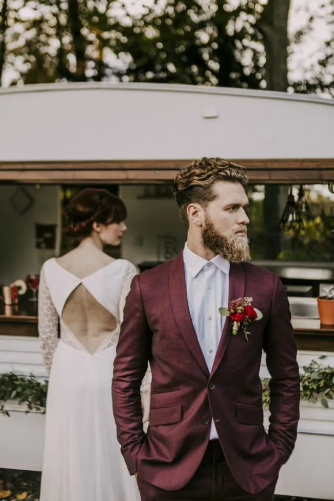 In this image for Mariage Elegant, Canada, showcasing the wedding tuxedo vendor Blandin & Delloye, a groom is dressed in a sophisticated deep burgundy tuxedo with a white dress shirt, adorned with a boutonniere featuring red and pink flowers. He stands confidently in front of a vintage-style mobile bar, which adds a rustic charm to the wedding setting. In the background, a bride in a white gown with a unique open back and delicate lace sleeves gazes away, enhancing the romantic atmosphere. The scene is set outdoors, under a canopy of trees, which adds a natural and serene backdrop to the elegant wedding attire display.