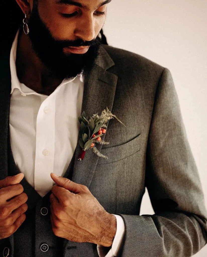 A groom adjusts his sleek, dark grey wedding suit jacket from Blandin & Delloye, fitting perfectly for a sophisticated wedding occasion. The suit features a subtle texture, complementing the crisp white shirt underneath. A small, elegant boutonniere composed of greenery and red berries is pinned to the lapel, adding a touch of color and style typical of Blandin & Delloye's offerings in the wedding tuxedo category. The groom's beard and long hair add to the overall elegant and modern aesthetic, suitable for an upscale wedding event in Canada.