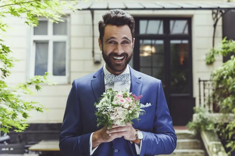 A groom stands outside, holding a small bouquet of white and pink flowers, in front of a classic building with black-framed windows and green foliage. He is wearing a stylish blue tuxedo with a patterned bow tie, exemplifying wedding attire from Blandin & Delloye in the Wedding Tuxedo category, highlighting sophistication and elegance perfect for a Canadian wedding setting.