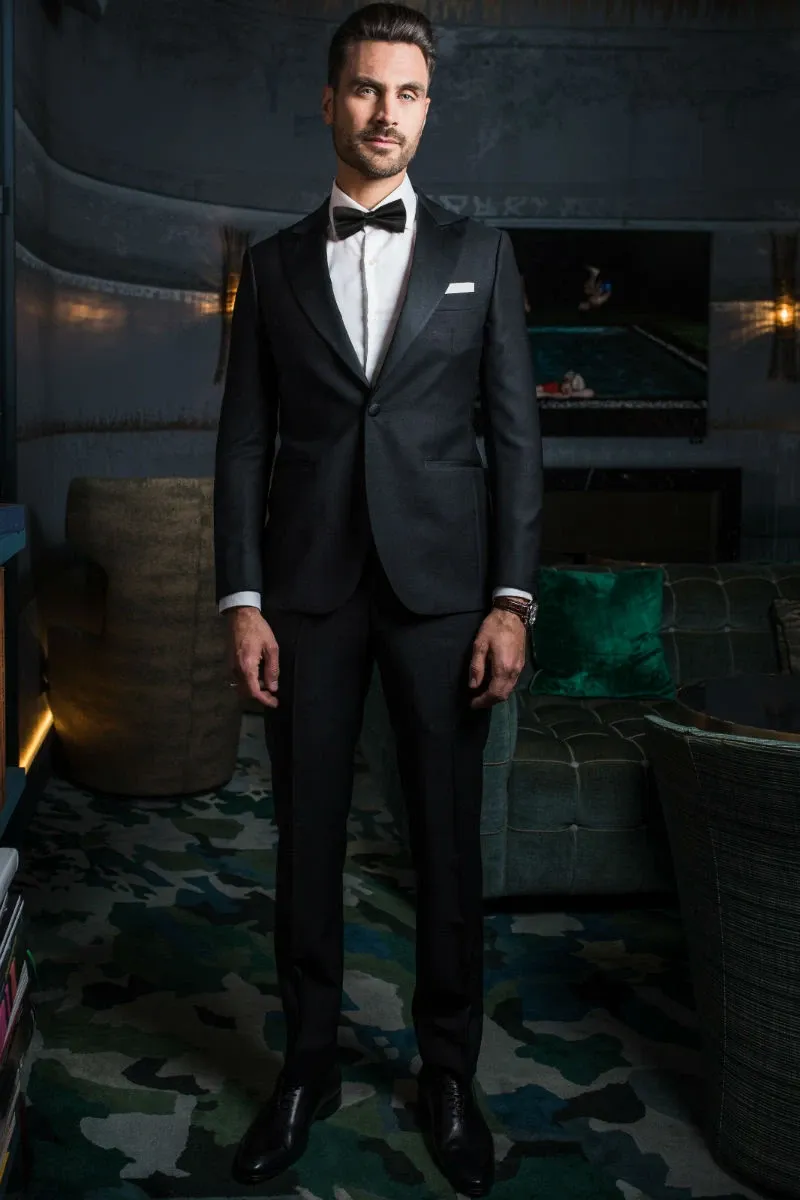 A groom is elegantly dressed in a classic black tuxedo designed by Blandin & Delloye, under the wedding tuxedo category for Mariage Elegant, Canada. The tuxedo features a sleek jacket with a satin lapel, a crisp white dress shirt, and a black bow tie, complemented by polished black shoes. The setting appears sophisticated, with dim lighting highlighting a modern, upscale interior that suggests a luxurious wedding venue or fitting room.