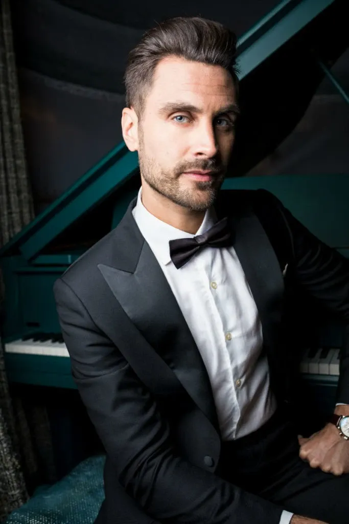 A groom is elegantly dressed in a classic black tuxedo by Blandin & Delloye, ideal for weddings. The tuxedo features a crisp white shirt and a stylish black bow tie. The groom is seated confidently in front of a grand piano, adding a touch of sophistication to the wedding attire display. This image is featured in the Wedding Tuxedo category for Mariage Elegant in Canada.