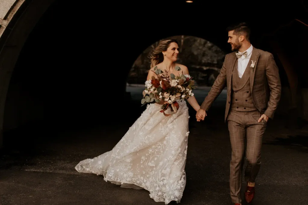A bride and groom walk hand in hand out of a dark tunnel, creating a striking contrast as they enter a sunlit area. The bride is elegantly dressed in a lace wedding gown with intricate floral detailing, holding a large bouquet of flowers. The groom, representing Blandin & Delloye in the wedding tuxedo category, is wearing a stylish brown plaid suit with a matching waistcoat and red shoes. His attire complements the sophisticated and modern theme of the wedding, as showcased on the Mariage Elegant, Canada platform. The couple's joyful expressions capture the essence of the celebration.