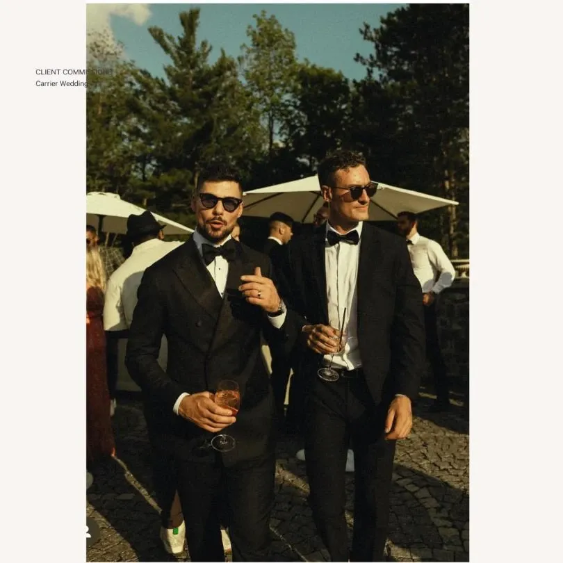 Two men are standing outdoors at a wedding event, dressed in elegant tuxedos from Maison Cloakroom, a vendor specializing in wedding tuxedos. They are both wearing sunglasses and holding glasses of rosé wine, adding a touch of sophistication to their attire. The setting appears festive, with other guests in the background and a large white umbrella providing shade. The scene captures the essence of a stylish wedding celebration, with a natural, sunlit ambiance surrounded by lush greenery.