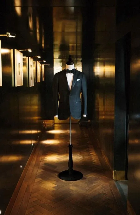 A sophisticated wedding tuxedo from Maison Cloakroom is elegantly displayed on a mannequin in a dimly lit hallway. The tuxedo features a sleek black jacket with a tailored fit, complemented by a classic white dress shirt and a stylish black bow tie. The polished wooden floor and ambient lighting highlight the luxurious fabric and meticulous design, reflecting the high-end quality of Maison Cloakroom's wedding attire. Frames on the wall add a touch of refinement to the setting, perfect for couples seeking elegant and timeless wedding tuxedos.