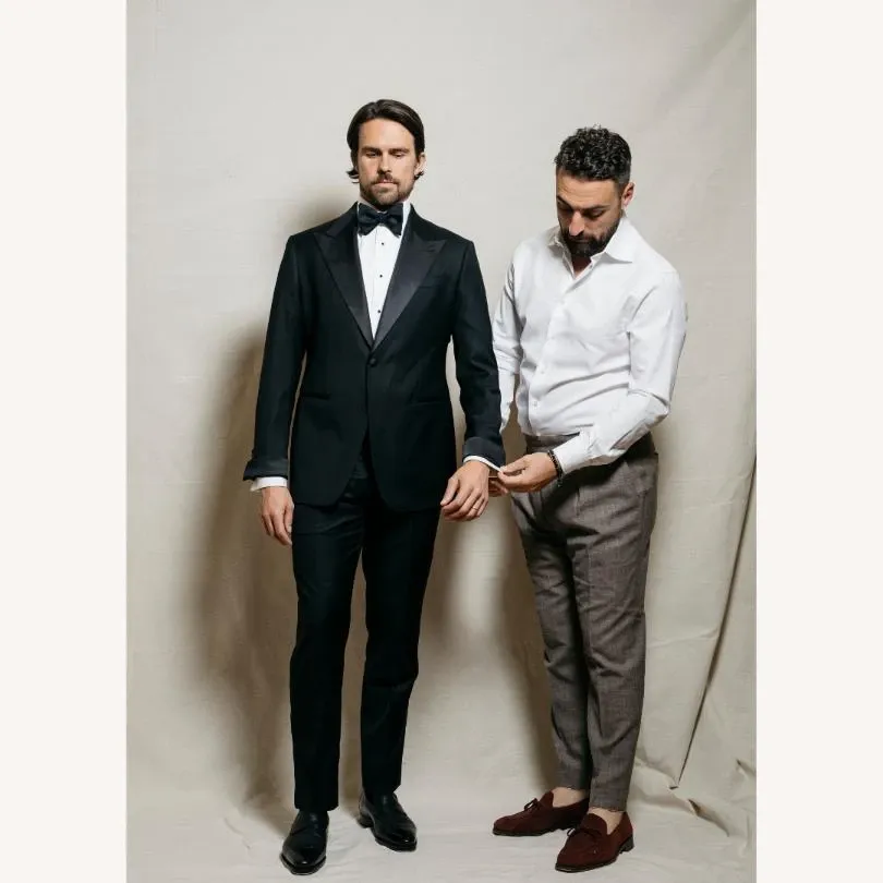 A well-dressed man is standing confidently in a black tuxedo with satin lapels, paired with a white dress shirt and a black bow tie. He is being attended to by another man, who is adjusting the tuxedo jacket. The second man is dressed in a white shirt and grey trousers, complemented by brown loafers. This image highlights the expertise of Maison Cloakroom in the Wedding Tuxedo category, showcasing their attention to detail and commitment to elegance, ideal for weddings. The setting has a simple, neutral backdrop, focusing attention on the refined tailoring and stylish attire suitable for a formal wedding.