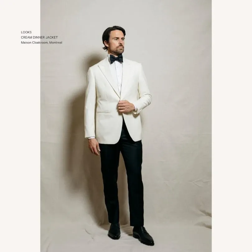 A man stands confidently in a sophisticated cream dinner jacket with a crisp white shirt and black bow tie, showcasing an elegant wedding tuxedo from Maison Cloakroom. The tailored jacket is complemented by black trousers and polished black shoes, ideal for a stylish wedding ensemble. The neutral backdrop enhances the refined and classic look, perfect for grooms seeking timeless elegance. This image represents the exquisite craftsmanship offered by Maison Cloakroom in the wedding tuxedo category for Mariage Elegant, Canada.
