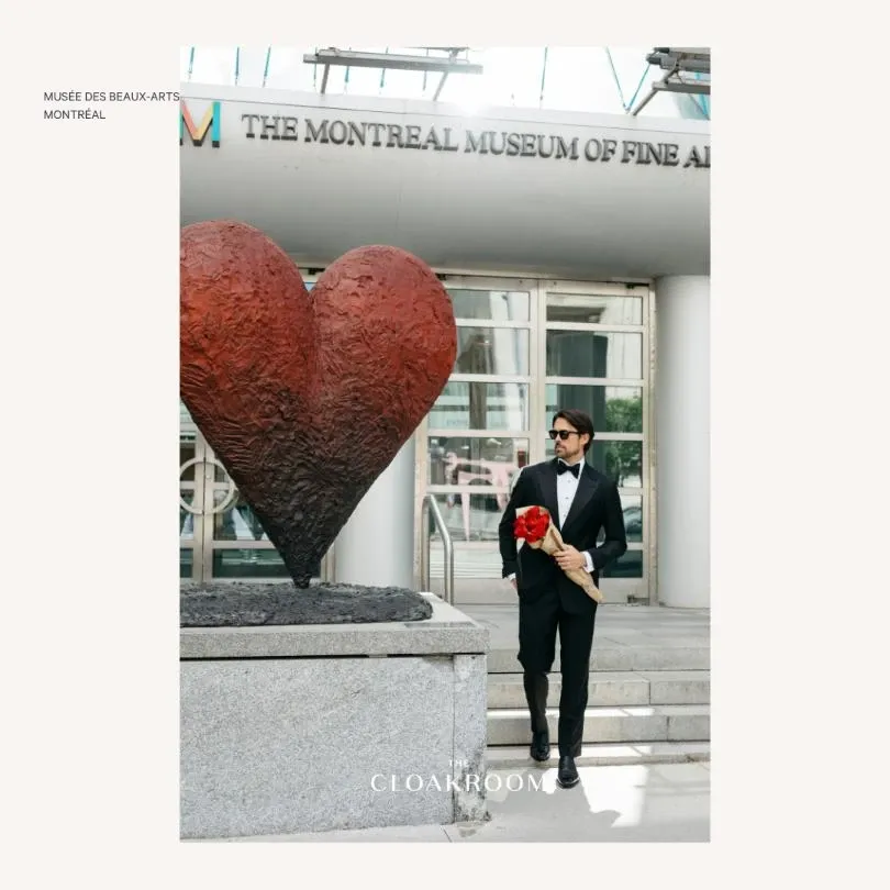 A groom wearing an elegant black tuxedo from Maison Cloakroom, a wedding tuxedo vendor, poses confidently at the entrance of the Montreal Museum of Fine Arts. He is holding a bouquet of vibrant red roses in one hand. The backdrop features a striking large red heart sculpture, adding a romantic touch to the scene. This setting provides a sophisticated and modern atmosphere, perfect for capturing a memorable wedding moment.