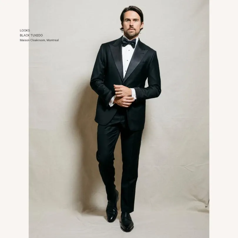 A well-dressed man is standing against a neutral background, wearing a classic black tuxedo from Maison Cloakroom, ideal for weddings. The tuxedo features a satin shawl lapel and is paired with a crisp white dress shirt and a black bow tie. The man has a poised and confident demeanor, perfectly showcasing the elegant attire designed for wedding ceremonies. His polished black dress shoes complement the formal ensemble, making this look suitable for a sophisticated wedding event. This image exemplifies the style and quality of wedding tuxedos offered by Maison Cloakroom.