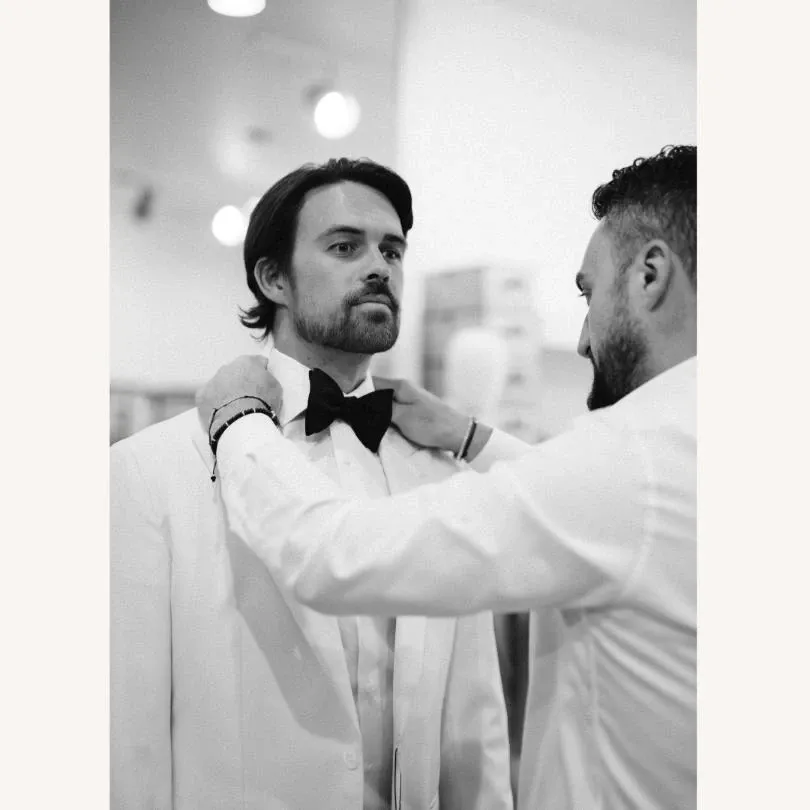 In this black and white image, a man is being fitted for a wedding tuxedo at Maison Cloakroom, a vendor under the Wedding Tuxedo category on Mariage Elegant, Canada. The gentleman, wearing a classic white tuxedo jacket and a black bow tie, is being assisted by a tailor who is adjusting his collar. The setting suggests a formal, elegant ambiance, typical for wedding preparation. The background exhibits soft lighting and blurred elements, highlighting the focus on the attire and the fitting process.