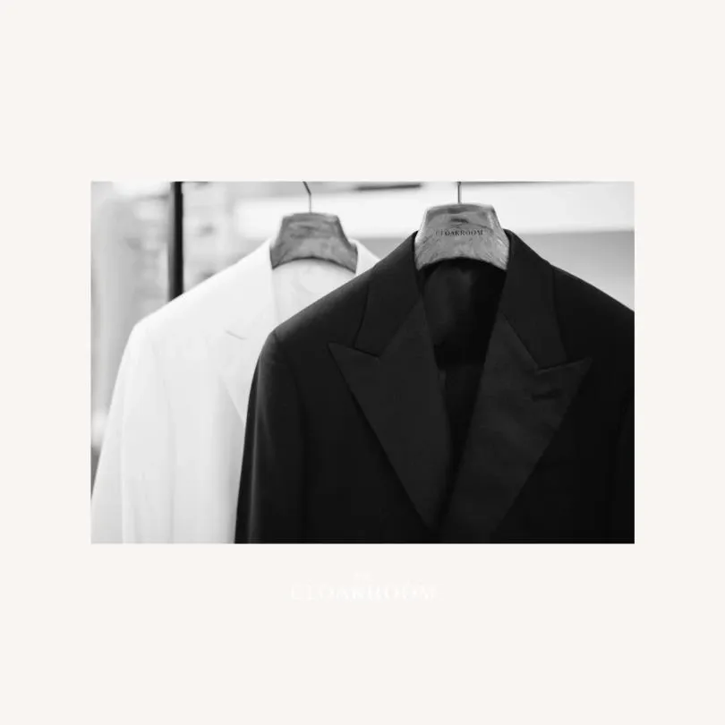 The image showcases two elegant tuxedos from Maison Cloakroom displayed on wooden hangers, ideal for weddings. The front tuxedo is a classic black with satin lapels, while the one behind it is white, reflecting a sophisticated choice for a wedding. Both tuxedos are positioned in a well-lit boutique setting, emphasizing the formal and stylish nature of wedding attire available from this vendor under the Wedding Tuxedo category.