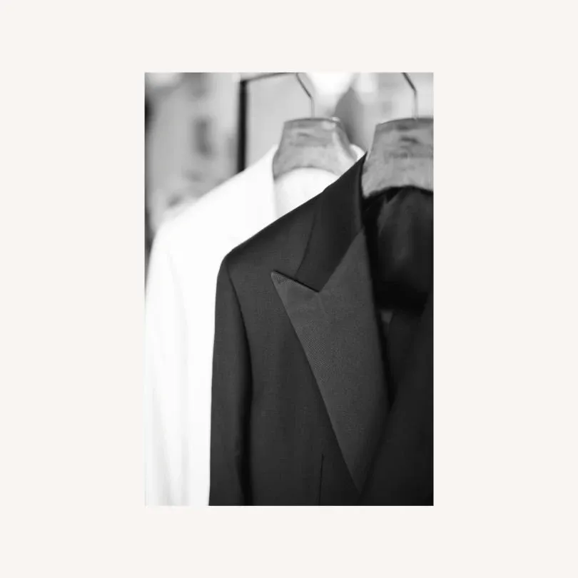 The image showcases a sophisticated black tuxedo jacket with sharp lapels, elegantly displayed on a wooden hanger in front of a blurred white garment. This tuxedo is part of the wedding collection from Maison Cloakroom, featured under the Wedding Tuxedo category on Mariage Elegant, Canada. The background is softly out of focus, emphasizing the exquisite tailoring and fabric texture of the tuxedo, ideal for a formal wedding setting.