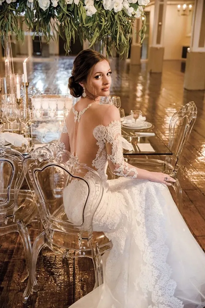 In this elegant image, a bride is seated at a formal dining table adorned with glassware and candles. She wears a stunning lace wedding dress designed by Charme Atelier, featured under the Wedding Dress category. The gown is intricately detailed with floral lace patterns and features a sophisticated open back design with delicate, off-the-shoulder lace sleeves. The setting is luxurious, with transparent chairs and a lush floral arrangement hanging overhead, providing a romantic atmosphere ideal for showcasing the refined craftsmanship of Charme Atelier's bridal fashion.