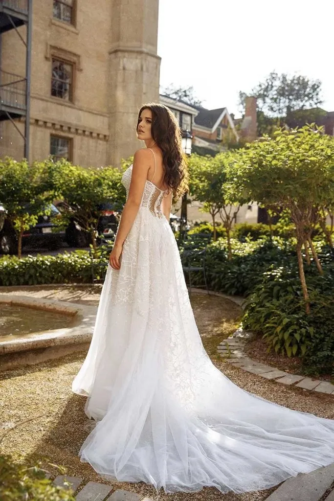 In the image from Mariage Elegant's vendor listing, Charme Atelier, a bride models an elegant wedding dress against a picturesque garden backdrop. The gown features a strapless design with a fitted bodice, adorned with intricate lace detailing, which flows seamlessly into a full, flowing train. The soft, ethereal fabric adds a touch of romance, complemented by the natural light highlighting its delicate textures. This stunning dress perfectly embodies the timeless elegance that Charme Atelier is renowned for in the wedding dress category.