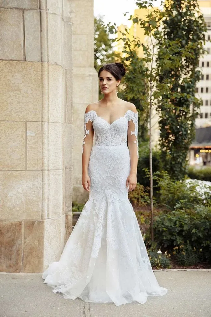 A bride stands elegantly outside, showcasing a stunning wedding dress from Charme Atelier in the wedding dress category of Mariage Elegant, Canada. The dress features intricate lace detailing with long, sheer lace sleeves and a fitted bodice that flows into a mermaid-style skirt. The off-the-shoulder neckline adds a touch of sophistication, highlighting the bride's poised stance. The background includes a stone building and lush greenery, offering a picturesque setting that complements the bridal attire perfectly.