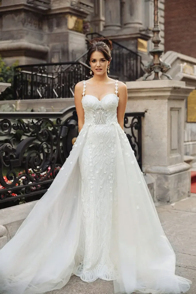 This image features a stunning wedding dress from the Charme Atelier collection, a vendor listed under the Wedding Dress category on Mariage Elegant, Canada. The bridal gown showcases a fitted silhouette with intricate lace detailing and elegant beaded straps. The dress is both sophisticated and modern, featuring a delicate train that adds a touch of grandeur. Set against an ornate wrought iron railing and classic stone architecture, the bride stands poised, highlighting the exquisite craftsmanship of Charme Atelier's design.