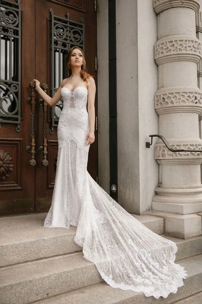 This image features a stunning wedding dress designed by Charme Atelier, displayed as part of their offerings in the wedding dress category on Mariage Elegant, Canada. The elegant gown has a form-fitting silhouette with intricate lace detailing from the bodice down to the flowing train. The bride stands gracefully on stone steps against a backdrop of ornate wooden doors and classical architecture, highlighting the grandeur and sophistication associated with Charme Atelier's bridal wear designs.