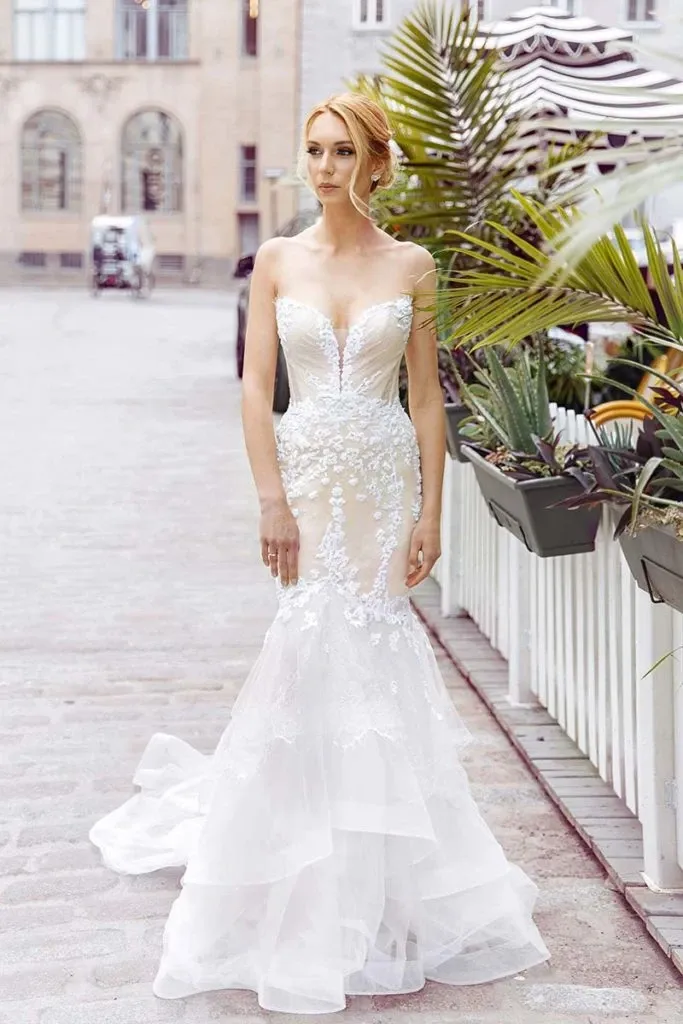A bride elegantly poses on a cobblestone street, wearing a stunning wedding dress by Charme Atelier, a featured vendor in the wedding dress category for Mariage Elegant, Canada. The gown showcases intricate lace detailing and a fitted mermaid silhouette that flows into a graceful train. The bride's hair is styled in an elegant updo, complementing the sophisticated and romantic look. The background features charming historic buildings and lush greenery, adding to the picturesque setting perfect for a wedding photoshoot.