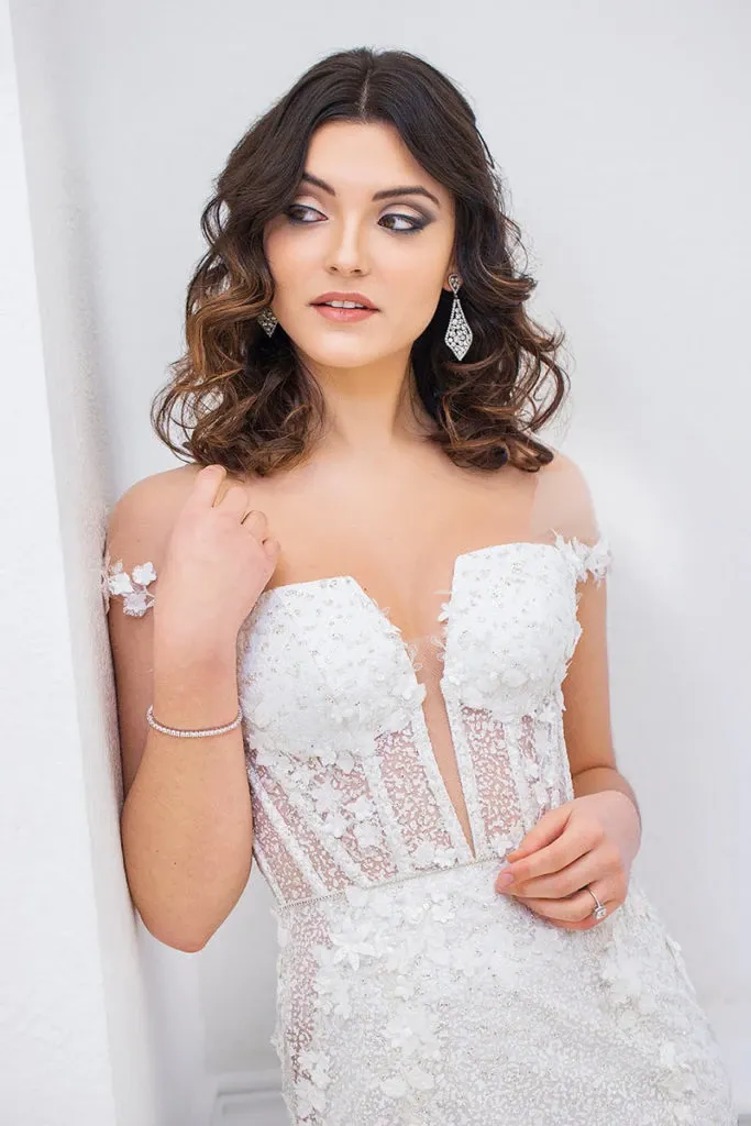 A bride stands gracefully in a luxurious wedding dress by Charme Atelier, showcasing the exquisite craftsmanship found in the Wedding Dress category on Mariage Elegant, Canada. The gown features a delicate lace bodice with intricate floral appliqués and a sheer panel elegantly highlighting the neckline. Her wavy brunette hair complements the elegant and sophisticated design, while sparkling drop earrings add a touch of glamour. The soft, light-hued backdrop enhances the dress's details, making it a perfect choice for a romantic wedding.