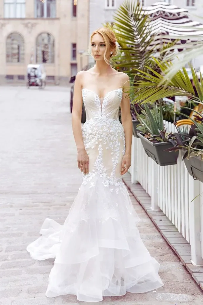A bride wearing a Charme Atelier wedding dress stands elegantly on a cobblestone street, exemplifying the essence of sophisticated wedding wear for Mariage Elegant in Canada. The gown features a fitted, strapless bodice adorned with intricate floral lace appliques that blend seamlessly into a flared, layered tulle skirt. Her hair is styled in a chic updo, complementing the overall elegant look. The backdrop includes charming urban architecture and lush green plants, creating a balance between modern style and natural beauty, ideal for a romantic wedding setting.
