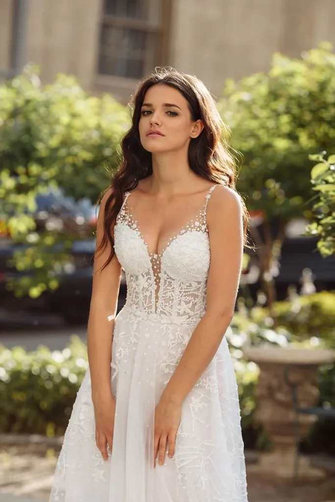 A bride stands outdoors wearing an elegant wedding dress by Charme Atelier, categorized under Wedding Dress at Mariage Elegant, Canada. The stunning dress features intricate lace detailing and a plunging neckline adorned with delicate floral embellishments. Its flowing, sheer fabric adds a touch of ethereal beauty, perfectly suited for a romantic garden wedding setting. The bride’s long, flowing hair complements the graceful design of the gown, enhancing the overall sophisticated look.