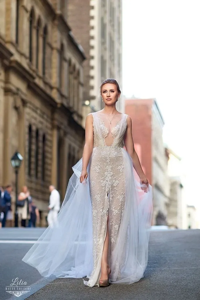 This image showcases a stunning wedding dress by Charme Atelier, fitting perfectly into the wedding dress category. The elegant gown features intricate lace detailing with a sheer overlay, creating a romantic and ethereal look. The dress is sleeveless with a deep V-neckline and a front slit, making it both modern and sophisticated. The bride is pictured walking confidently down a city street, with historic buildings blurred in the background, highlighting an urban fairytale wedding theme. This visually striking composition emphasizes the blend of contemporary and classic bridal fashion.