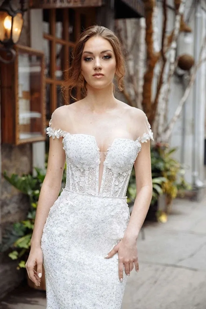 The image features a bride wearing an elegant wedding dress by Charme Atelier, a prominent vendor in the wedding dress category for Mariage Elegant, Canada. The dress is a stunning off-the-shoulder design with delicate lace and floral embellishments, creating a romantic and sophisticated look. The bodice is form-fitting with intricate detailing, and the skirt flows gracefully, complemented by a natural, outdoor background with rustic elements. This setting enhances the timeless elegance of the wedding dress, making it suitable for brides seeking classic and refined bridal wear.
