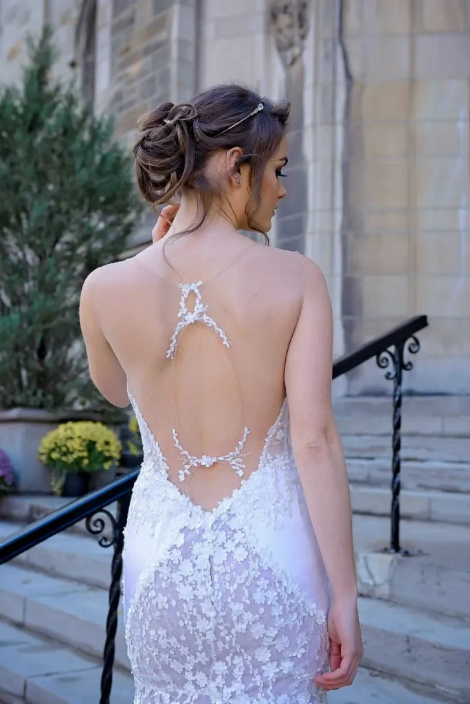 This image showcases a stunning wedding dress designed by Charme Atelier, featured in the wedding dress category on Mariage Elegant, Canada. The gown elegantly displays a backless design with intricate lace detailing extending from the shoulders down to the lower back. The dress features delicate floral lace patterns that cascade gracefully, adding an ethereal touch. The bride's hair is styled in a sophisticated updo, highlighting the exquisite details of the dress. The setting includes a stone church entrance with steps and subtle floral decorations, providing a classic and romantic wedding atmosphere.