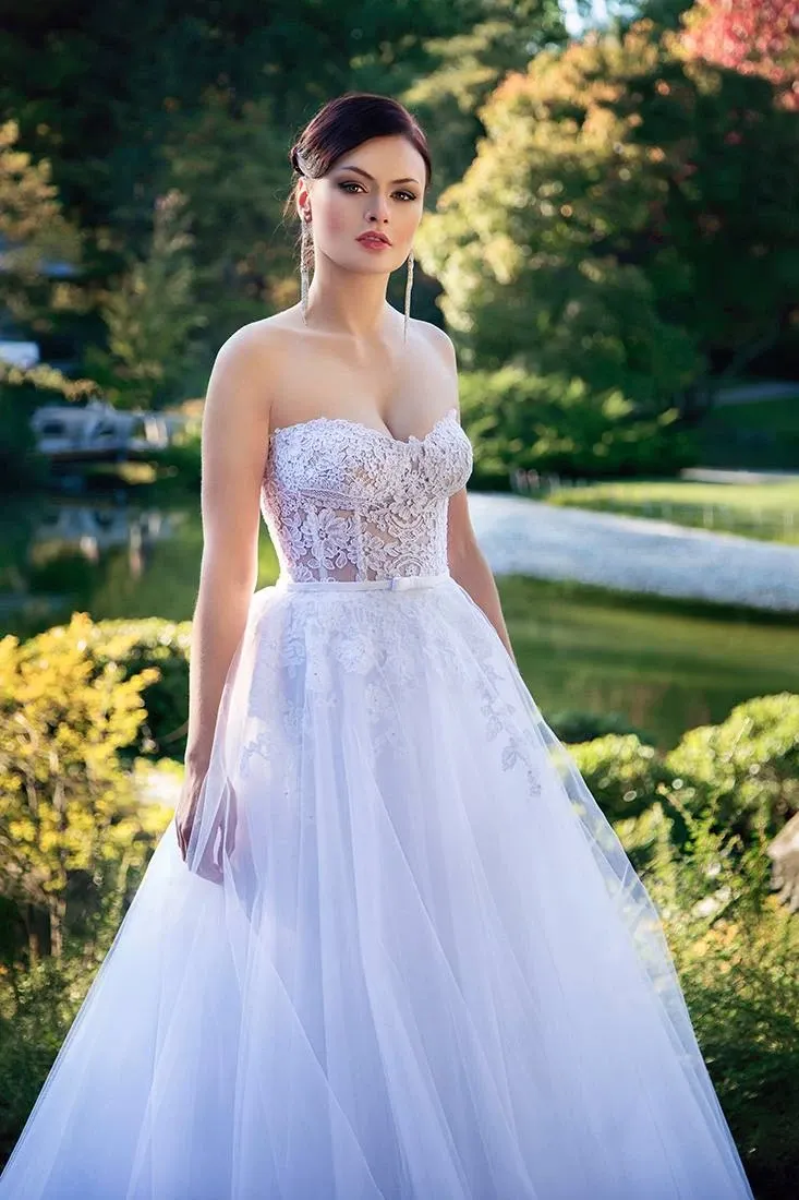 This image showcases a stunning wedding dress by Charme Atelier, featured in the Wedding Dress category on Mariage Elegant, Canada. The gown is an exquisite strapless design with a fitted lace bodice, offering delicate floral details and elegant embroidery. Its voluminous tulle skirt flows gracefully, creating a romantic and timeless silhouette. The bride stands outdoors, surrounded by lush greenery and a serene pond, adding a picturesque backdrop to the bridal look.