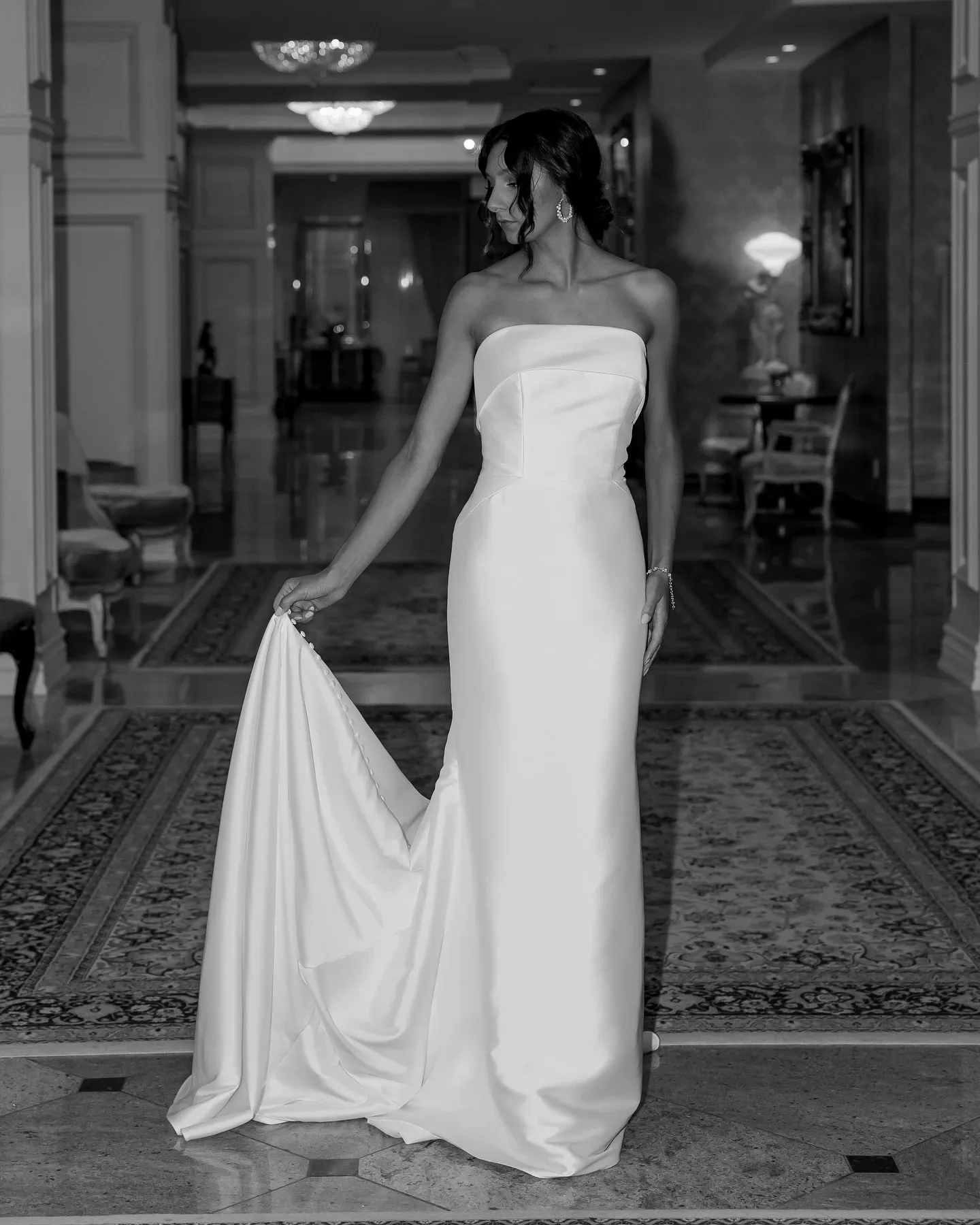 A bride is elegantly posing in a luxurious hallway, wearing a strapless, floor-length wedding gown by La Jolie Robe, a vendor in the Wedding Dress category on Mariage Elegant, Canada. The gown features a sleek, fitted silhouette with a straight neckline and a flowing train that she delicately holds with one hand. The setting is opulent, with plush chairs, ornate rugs, and warm lighting, highlighting the sophisticated ambiance of a high-end venue. Her hair is styled in an elegant updo, complemented by dangling earrings, adding to the overall sophisticated bridal look.