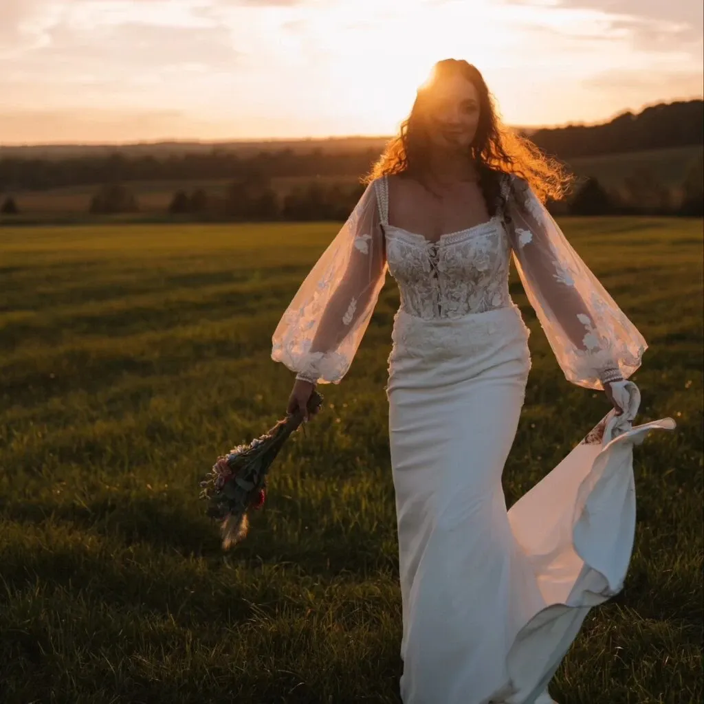 In this image from Mariage Elegant's wedding planning site, a model showcases a stunning wedding dress by the vendor La Jolie Robe. The gown features intricate lace detailing on the bodice and elegant, sheer puff sleeves adorned with floral embroidery. The simple yet sophisticated skirt flows gracefully, enhancing the romantic aesthetic. The model is holding a bouquet and walking through a lush, sunlit field, creating a picturesque and serene wedding setting.