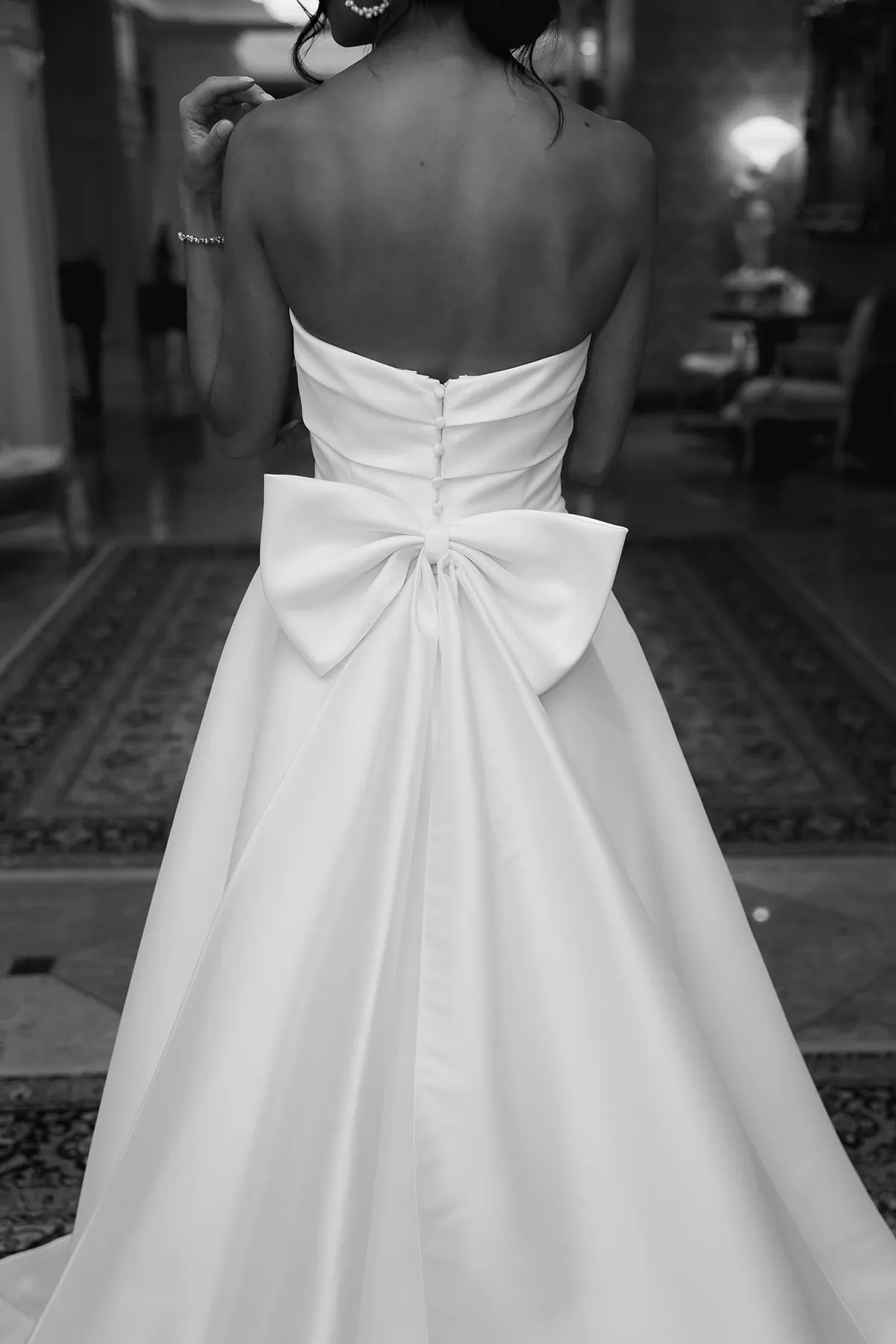 This elegant image showcases a stunning strapless wedding dress by La Jolie Robe, a featured vendor in the wedding dress category on Mariage Elegant, Canada. The gown is expertly tailored, with a smooth satin finish and a fitted bodice. The back of the dress is adorned with a large, statement bow, adding a touch of sophistication and charm. The scene is set in a luxurious setting with plush carpets and ornate furnishings, enhancing the overall atmosphere of elegance and refinement, ideal for a grand wedding celebration.
