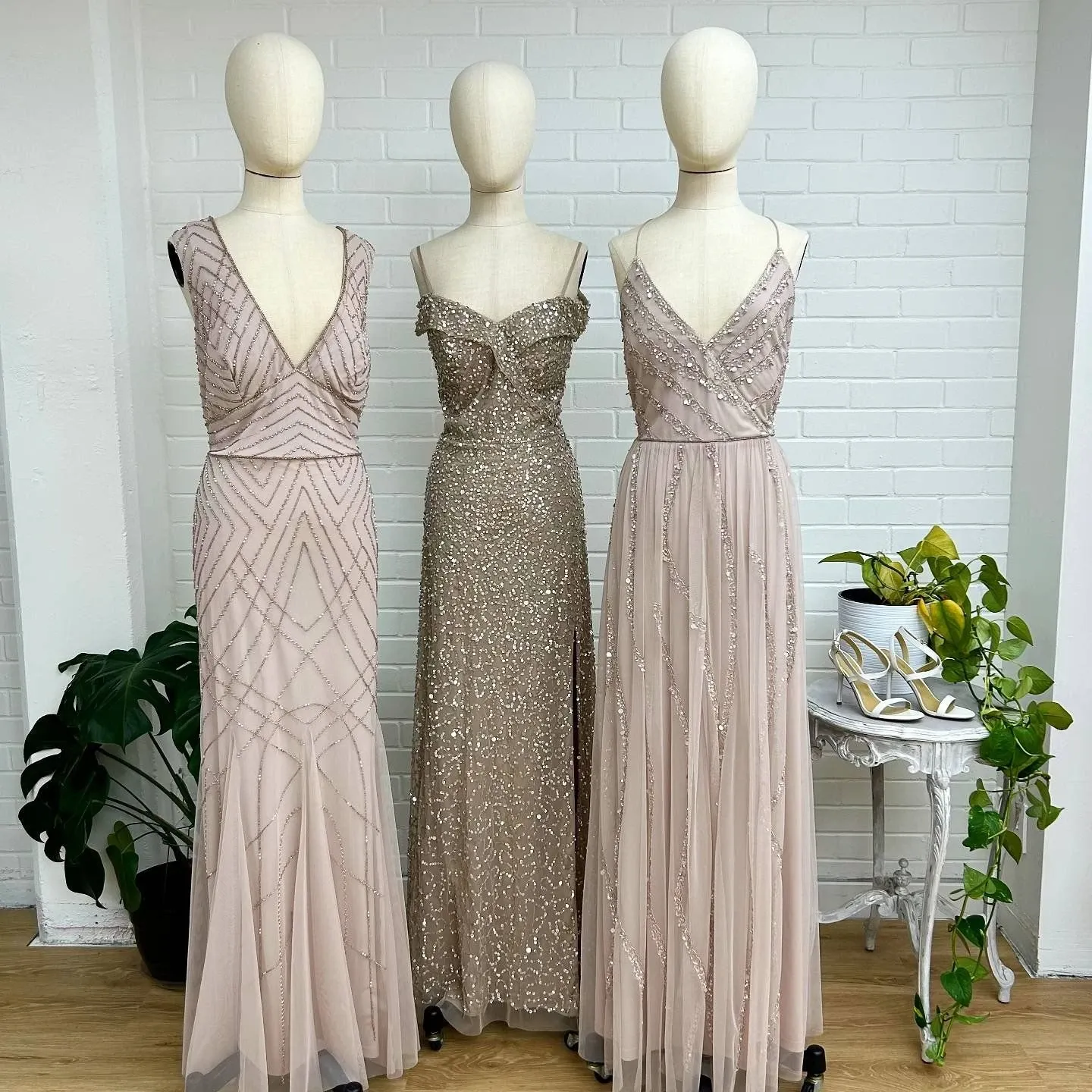 Three elegant wedding dresses from the vendor La Jolie Robe are displayed on mannequins against a white brick wall. Each dress is intricately designed, showcasing the beauty of the wedding dress category. The dress on the left features a plunging neckline with geometric bead patterns. The center dress is adorned with shimmering embellishments and has an off-the-shoulder design. The dress on the right, with delicate spaghetti straps, flows gracefully with a tulle overlay and subtle sequin detail. A potted plant and a small white table with a pair of heels add to the sophisticated setting, emphasizing the attention to detail provided by La Jolie Robe in creating exquisite wedding attire.