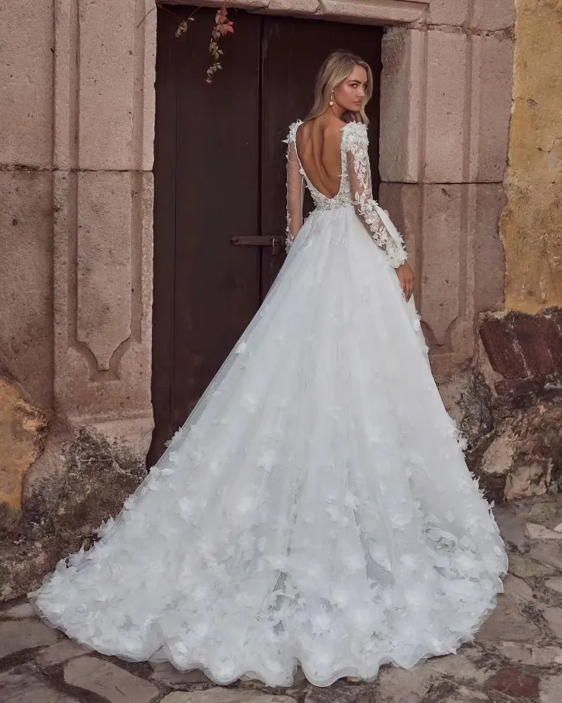 In this elegant image for Mariage Elegant's wedding planning site, the vendor La Jolie Robe showcases a stunning wedding dress. The dress features a full, flowing skirt adorned with intricate floral appliqués, adding texture and grace. The fitted bodice has long sleeves and delicate lace detailing, beautifully complementing the deep, open back design. The bride stands in front of a rustic stone building, enhancing the romantic atmosphere perfect for a wedding. This image highlights the exquisite craftsmanship and timeless style La Jolie Robe offers in their wedding dress collection.