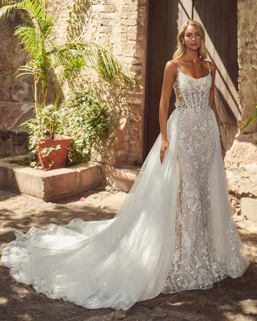 This image features a stunning wedding dress by La Jolie Robe, a vendor in the wedding category on Mariage Elegant, Canada. The dress is elegantly designed with intricate lace detailing extending from the bodice down to the flowing train. Its semi-sheer bodice adds a modern touch, complemented by thin shoulder straps. The dress stands out beautifully against a rustic stone wall backdrop, with lush green plants adding a natural element to the setting. The scene captures a sense of elegance and romance, perfect for a memorable wedding day.