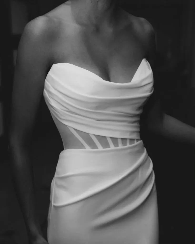 The image features a model wearing an elegant wedding dress by La Jolie Robe, part of the wedding dress category. The dress showcases a strapless design with a beautifully draped bodice that elegantly accentuates the waist. It features subtle, sheer detailing at the sides, adding a modern touch to the classic silhouette. This sophisticated gown epitomizes the refined and stylish choices available for brides planning their weddings through Mariage Elegant in Canada.