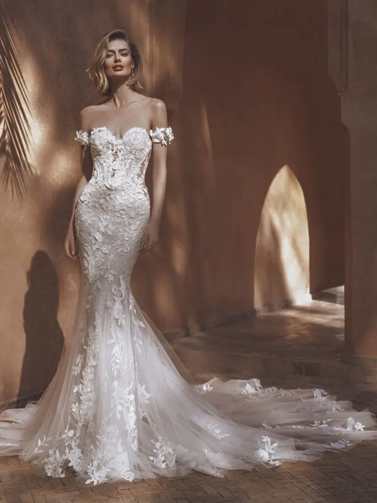 The image features a stunning wedding dress from La Jolie Robe, showcased in the context of wedding dress planning under the vendor category. The gown is an off-the-shoulder design with intricate floral lace details covering the bodice and flowing elegantly into a sheer, fitted skirt. The dress includes delicate floral appliques placed strategically along its length, adding a touch of romantic elegance. The bride stands in a softly lit, rustic setting with a warm, earthy background that enhances the gown's delicate beauty and sophistication, perfect for a high-class, elegant wedding ceremony.