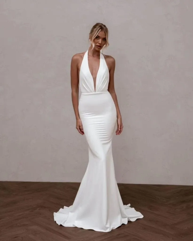 A model elegantly poses in a sleek, form-fitting wedding dress by La Jolie Robe, featured in the Wedding Dress category of Mariage Elegant, Canada. The gown is a stunning white with a deep V-neck halter that accentuates the model's neckline and shoulders. It flows gracefully to the floor, creating a sophisticated and timeless silhouette against a minimalist studio backdrop.