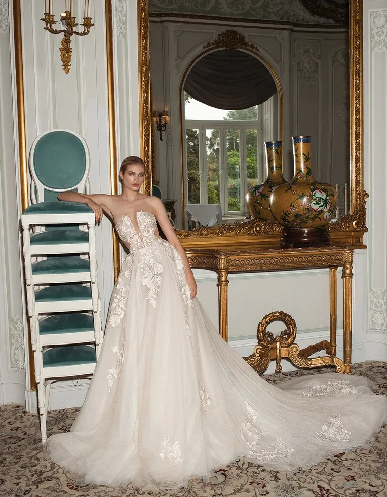 This image showcases a stunning wedding dress from White Montreal, categorized under Wedding Dress on Mariage Elegant. The elegant gown features intricate lace appliqués and a voluminous tulle skirt, exuding a sense of timeless sophistication. The model, poised in a luxurious setting, enhances the grandeur of the dress. Graceful detailing and a strapless neckline complete the look, making it an exquisite choice for brides seeking elegance. The opulent backdrop includes ornate wall decorations, a large mirror with an ornate gold frame, and plush classical furniture, amplifying the dress's majestic appearance.