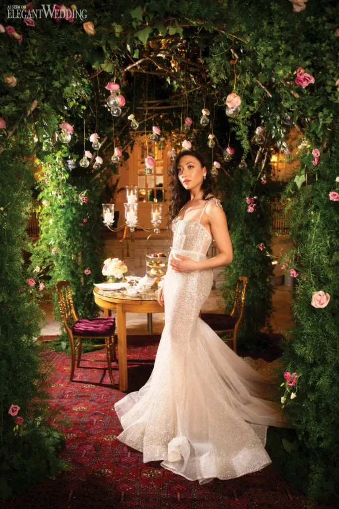 A bride wearing an elegant, form-fitting wedding dress by White Montreal stands gracefully beneath a beautiful green archway adorned with lush foliage and delicate pink roses. The gown, classified under the Wedding Dress category, features sparkling fabric with thin shoulder straps and a trumpet silhouette that flares at the bottom. The setting, suitable for a sophisticated wedding, includes hanging glass candle holders and a small table with ornate decorations, creating a romantic and enchanting atmosphere. The backdrop suggests a luxurious venue, ideal for the perfect wedding celebration with Mariage Elegant, Canada.